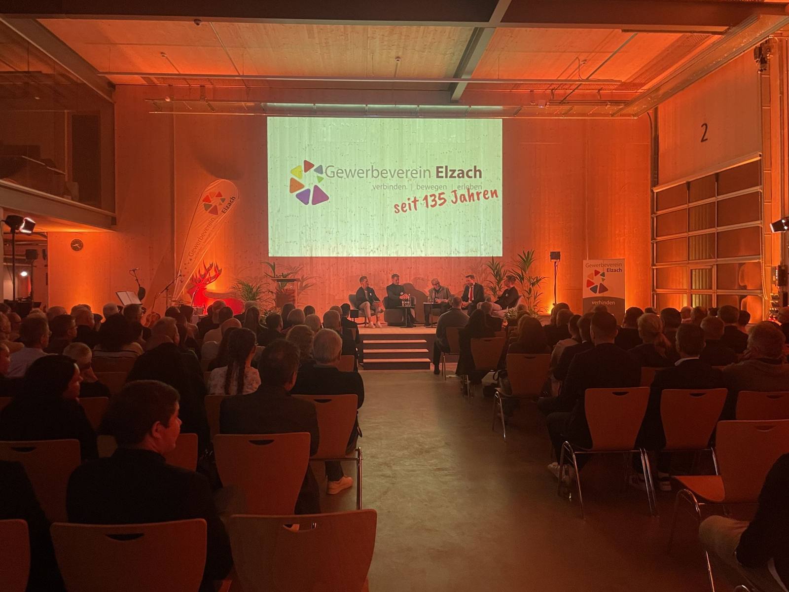 Talkrunde auf der Bühne vor großer Projektion, Saal in warmem Licht