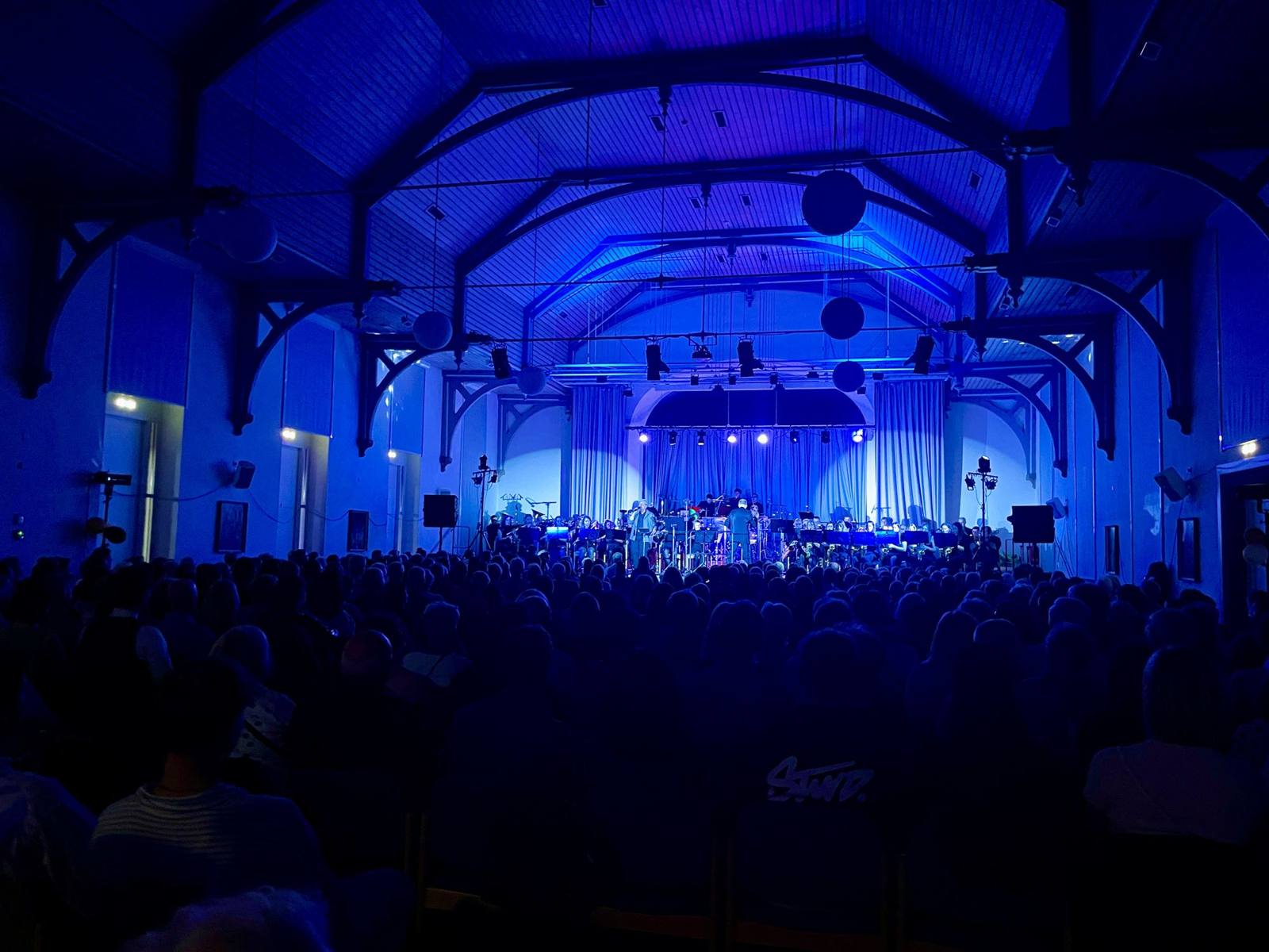 Blau beleuchtete Halle mit Orchester auf der Bühne und vollem Saal